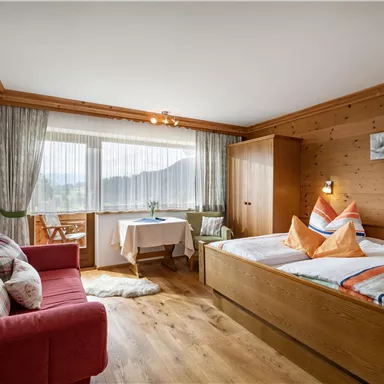 A cozy bedroom with wooden paneling and large windows. There is a double bed, a comfortable seating area, and a table with a view of the mountains.