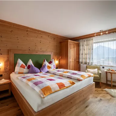 A cozy bedroom with a large double bed and colorful cushions. The walls are made of wood, and there is a window with a view of the mountains.
