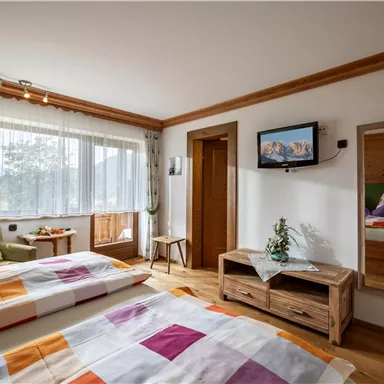 A cozy bedroom with two beds and colorful bed covers. The room has large windows, a television, and a modern wooden dresser.