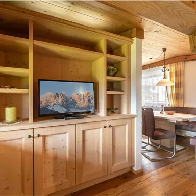 A cozy interior with wooden shelves and a television. In the background, there is a dining area with chairs and a table.