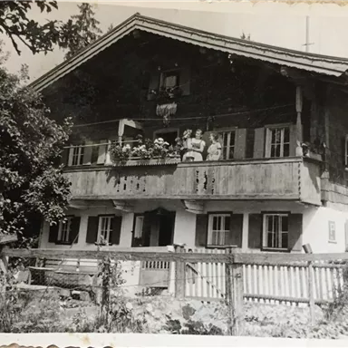 A traditional old house with a balcony and blooming plants. In front of the house, there are several people, and it is surrounded by trees.