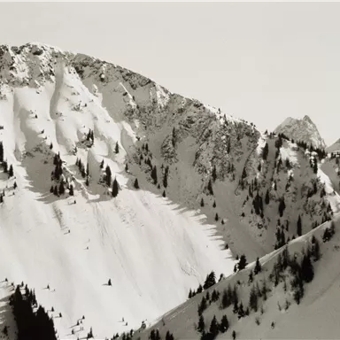 A snowy mountain landscape with high peaks and some green coniferous trees. The sky appears clear and bright.