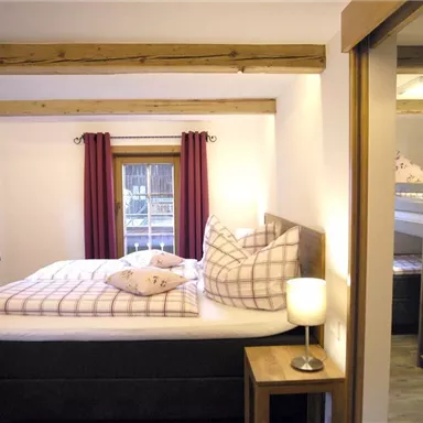 A cozy bedroom with a large bed and checkered pillows. The windows are decorated with red curtains and a bedside table with a lamp is also present.