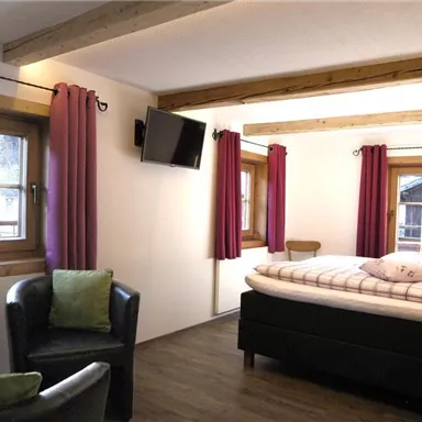 A cozy room with a large bed and two windows. The walls are painted in neutral colors and decorated with red curtains.