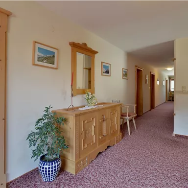A bright hallway with a wooden table and decorative pictures on the wall. The floor is covered with a red carpet, and at the end of the hallway, more doors are visible.