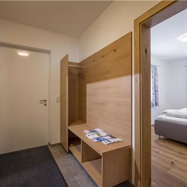 A bright hallway with wooden details and a bench. A view into an adjacent room with a bed and windows.