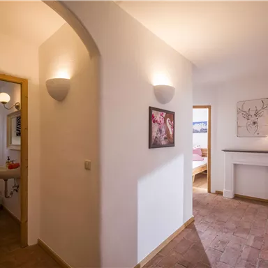 A bright hallway with white walls and warm light. You can see an entrance to a bathroom and a view of a cozy living room.