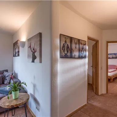 A bright hallway with a cozy seating area and pictures on the walls. Through a door, a bedroom can be seen.