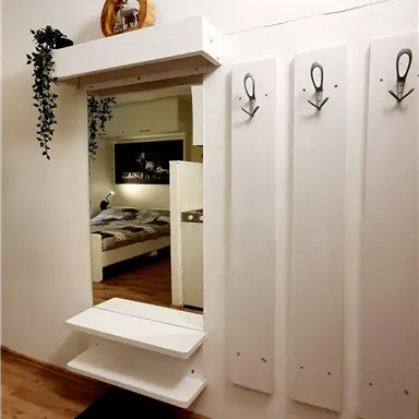 A modern entrance area with a white mirror and coat hooks. In the background, a bedroom with a double bed is visible.