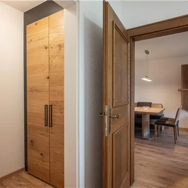 A modern entrance area with a wooden cabinet and a mirror. In the background, there is a dining area with a table and chairs visible.