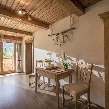 Ein helles, gemütliches Zimmer mit Holzboden und einem Tisch mit Stühlen. Große Fenster bieten einen Blick auf die Natur und sorgen für viel Licht.