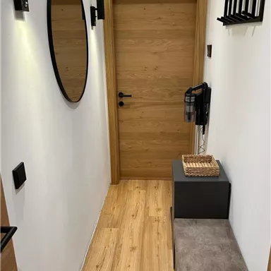 Ein schmaler Flur mit einem Holzfußboden und einer hellen Wandfarbe. Es gibt einen Spiegel, eine Eingangstür und eine kleine Sitzgelegenheit mit einem Ablagetisch.