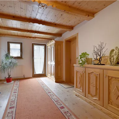 A bright hallway with wooden ceilings and a carpet. On the left, there is a decorative cabinet and on the right, a dresser with plants.