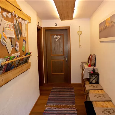 Ein einladender Flur mit Holzelementen und einer Pinnwand voller Informationen. Rechts stehen ein Tisch mit Dekoration und einige Sitzgelegenheiten.