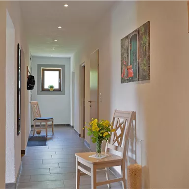 Ein heller Flur mit weißen Wänden und grauem Boden. Auf der linken Seite stehen Sitzgelegenheiten und ein Tisch mit einer Blumenvase.