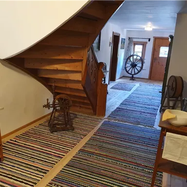 An inviting hallway with a wooden staircase and a colorful rug. On the left side, there is an antique piece of furniture and a spinning wheel.