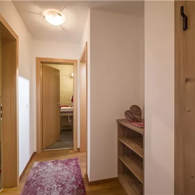 Ein heller Flur mit Holztüren und einem Holzregal. Am Boden liegt ein Teppich und im Hintergrund ist ein Badezimmer zu sehen.