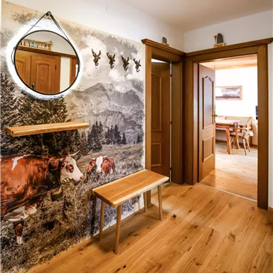 Ein einladender Flur mit einem Holzboden und einem Spiegel in Tropfenform. An der Wand ist ein freundliches Alpenmotiv mit Kühen und Bergen zu sehen.