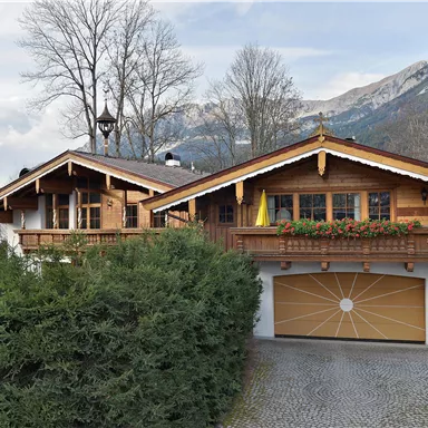 A charming wooden house in alpine style, surrounded by trees. In the background, majestic mountains and a cloudy sky can be seen.