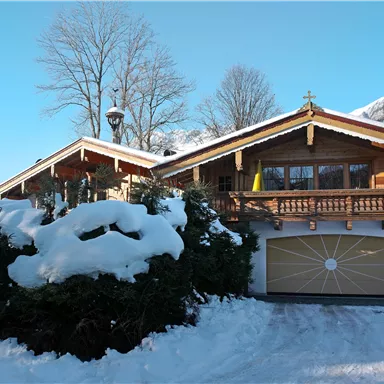A beautiful wooden house in the snow with a large balcony. Surrounded by snow-covered trees and a clear blue sky.