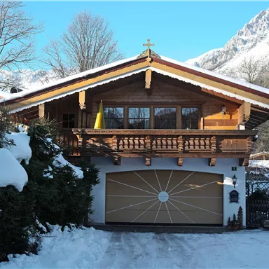 A charming wooden house with a balcony, surrounded by snow-covered trees. In the background, mountains are visible under a clear blue sky.