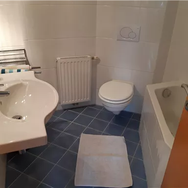 A modern bathroom with a sink, toilet, and bathtub. The bright room has gray tiles and an orange door.