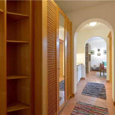 Ein heller Flur mit einem Holzschrank und einem Teppich. Im Hintergrund ist ein weiteres Zimmer mit einem Stuhl und Bilderrahmen zu sehen.