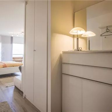 A modern bedroom with a white wardrobe and a stylish sideboard. The room is well-lit and has large windows.