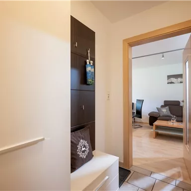 Ein heller Flur mit einem Schuhschrank und einer offenen Tür zum Wohnzimmer. Im Hintergrund sind ein Sofa und ein Tisch sichtbar.