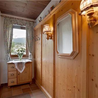 A cozy hallway with wooden-paneled walls and a large window. On the dresser sits a flower vase, and the wall lights provide a gentle glow.