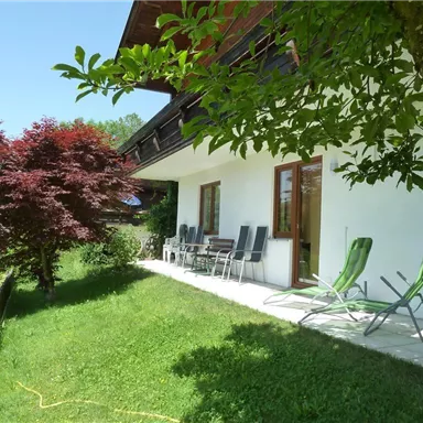 Ein gemütlicher Außenbereich eines Hauses mit Blick auf einen gepflegten Garten. Entspannte Liegen und ein roter Baum schaffen eine einladende Atmosphäre.