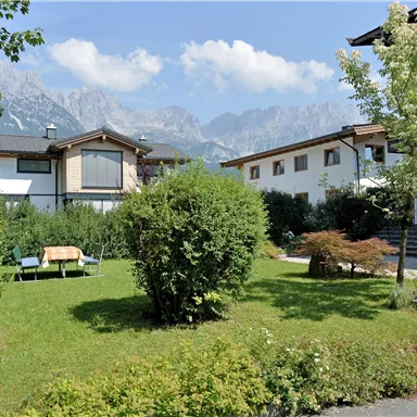 Ein schöner Garten mit gepflegtem Rasen und einigen Bäumen. Im Hintergrund sind Berge und moderne Häuser zu sehen.