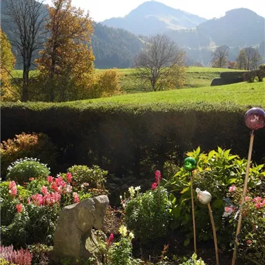 Ein schöner Garten mit bunten Blumen und einer Steinskulptur im Vordergrund. Im Hintergrund sind sanfte Hügel und Bäume vor einem klaren Himmel zu sehen.