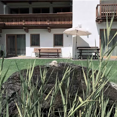 Ein ruhiger Garten mit einer Steinfontäne und üppigem Grün. Im Hintergrund ist ein modernes Gebäude mit Balkonen und einem Sonnenschirm.