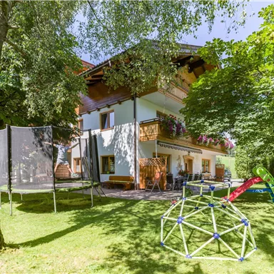 Ein schönes Haus umgeben von Bäumen und einem Garten. Im Vordergrund befinden sich eine Trampolin und ein Spielgerät für Kinder.