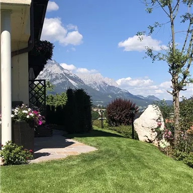 A picturesque view of mountains under a clear blue sky. In the foreground, there is a well-maintained garden with flowers and green spaces.
