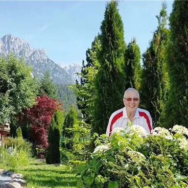 A cheerful man stands in a garden with tall, green trees. In the background, impressive mountains and a blue sky are visible.
