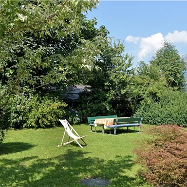Ein schöner Garten mit grünem Rasen, Bäumen und einem Tisch mit Bänken. Im Vordergrund steht eine Liege und im Hintergrund sind Berge zu sehen.