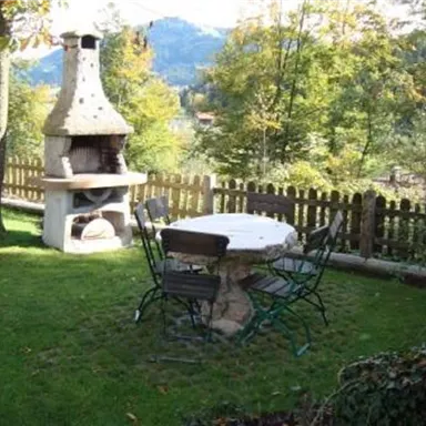A cozy garden with a grill and a round table. Green grass and trees surround the seating area.