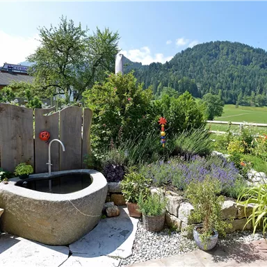 Ein schöner Garten mit Blumen und einem stillen Wasserbecken. Im Hintergrund sind grüne Hügel und ein strahlend blauer Himmel zu sehen.