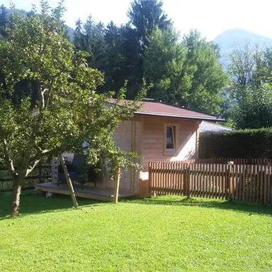 A beautiful garden with a wooden house and a lush green lawn. In the background, trees and mountains can be seen.