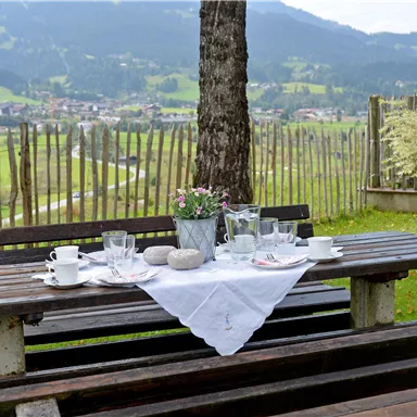 Ein schöner Tisch im Freien, gedeckt mit Geschirr und einer Blumenmitte. Im Hintergrund sind sanfte Hügel und eine malerische Landschaft zu sehen.
