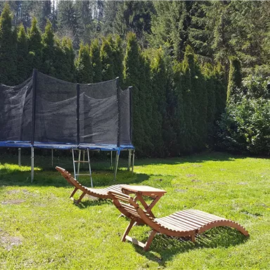 Ein weitläufiger Garten mit einer Trampolin und Liegen. Umgeben von hohen Hecken und Bäumen.