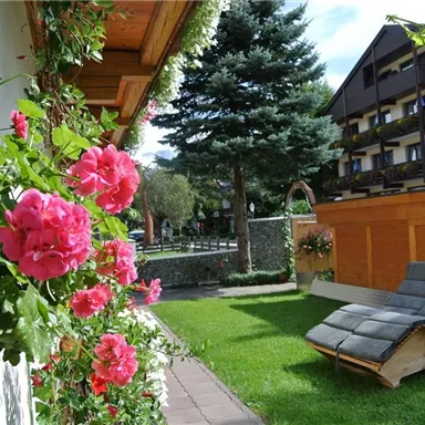 A beautiful garden with colorful flowers and a deck chair. In the background, trees and a building can be seen.