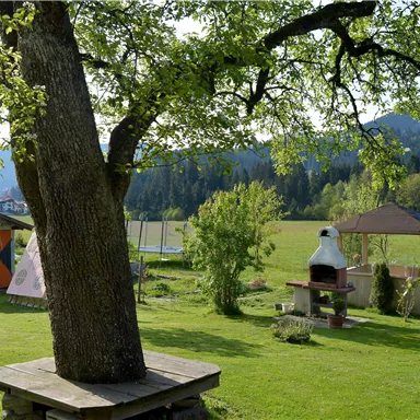 A beautiful garden with a large tree, a cozy garden gazebo, and a grilling area. In the background, there are meadows and gentle hills visible.