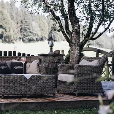 Eine gemütliche Terrasse mit einem rattanmöbel Set und Kissen. Im Hintergrund sind grüne Wiesen und Bäume zu sehen.