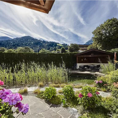 Ein schöner Garten mit farbenfrohen Blumen und einem kleinen Teich. Im Hintergrund sind sanfte Hügel und eine klare, bewölkte Himmel zu sehen.