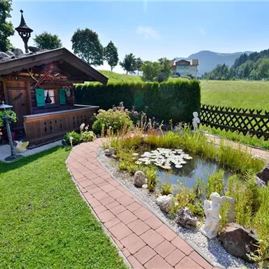 Ein gemütliches Gartenhaus steht in einem schönen, grünen Garten. Neben dem Gartenhaus befindet sich ein Teich mit Seerosen und üppigen Pflanzen.