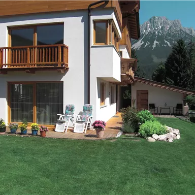 Ein modernes Haus mit einem gepflegten Garten und gemütlichen Liegen auf der Terrasse. Im Hintergrund sind majestätische Berge zu sehen.