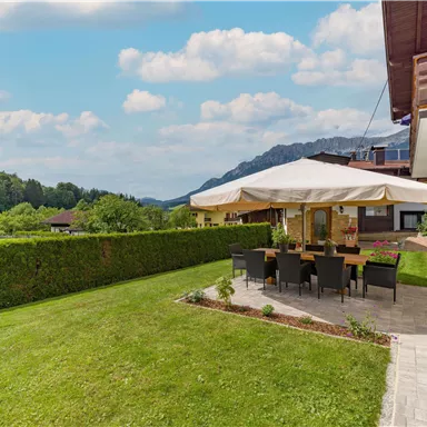 A terrace with a large sunshade and an outdoor dining area. The view overlooks a green meadow and mountains in the background.